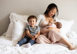mother breastfeeding newborn on bed in pink dress with toddler sitting next to her. 