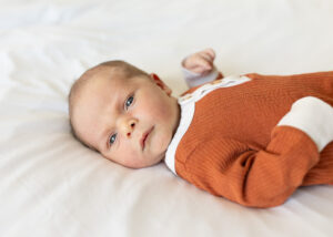 Awake newborn laying on white sheet looking at camera in a burnt orange romper. 