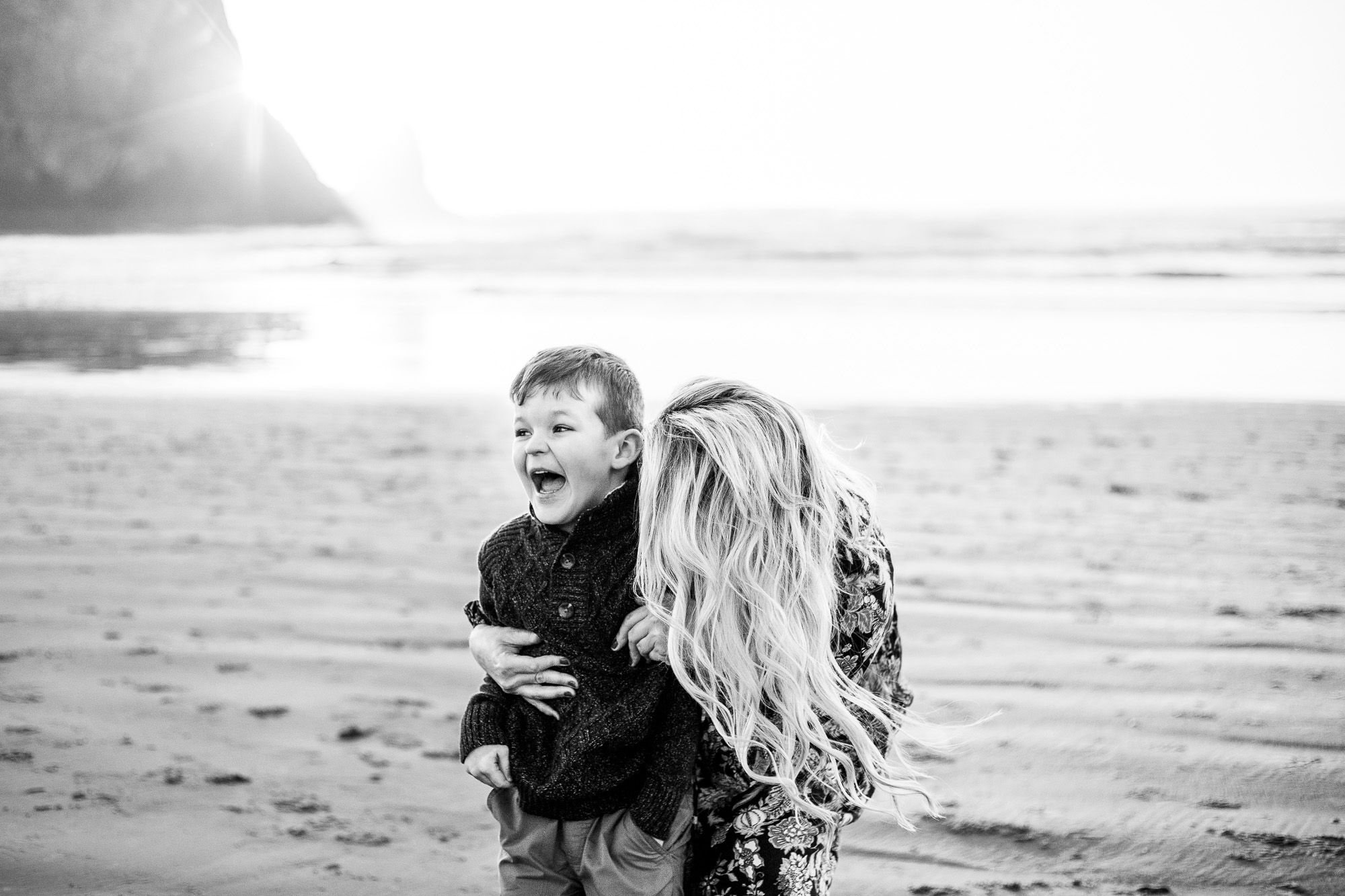 Black and white image of mom hugging son while he laughs at beach