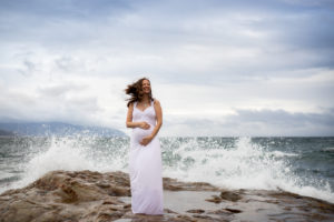 Pregnant woman holding belly with waves crashing behind her at Cape Kiwanda State Park