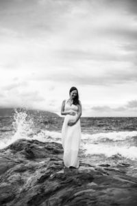 black and white image of pregnant woman with waves crashing behind her on the beach at Cape Kiwanda State Park