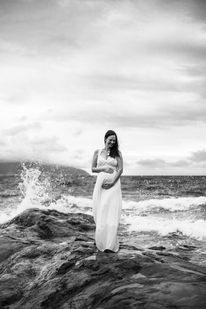 A pregnant woman standing on rocks in Pacific City with waves crashing behind her. | Oregon Coast Family Photographer 
