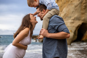 A family of three on the beach looking at one another laughing while mom is holding pregnant belly. Pacific City, Oregon Hotels. 