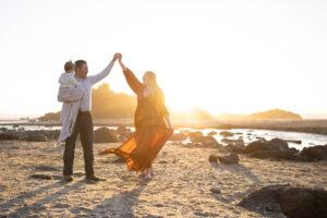 Couple dancing on beach at sunset while dad is holding infant. 