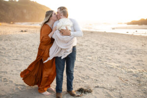 Couple kissing on beach at sunset while holding infant. 