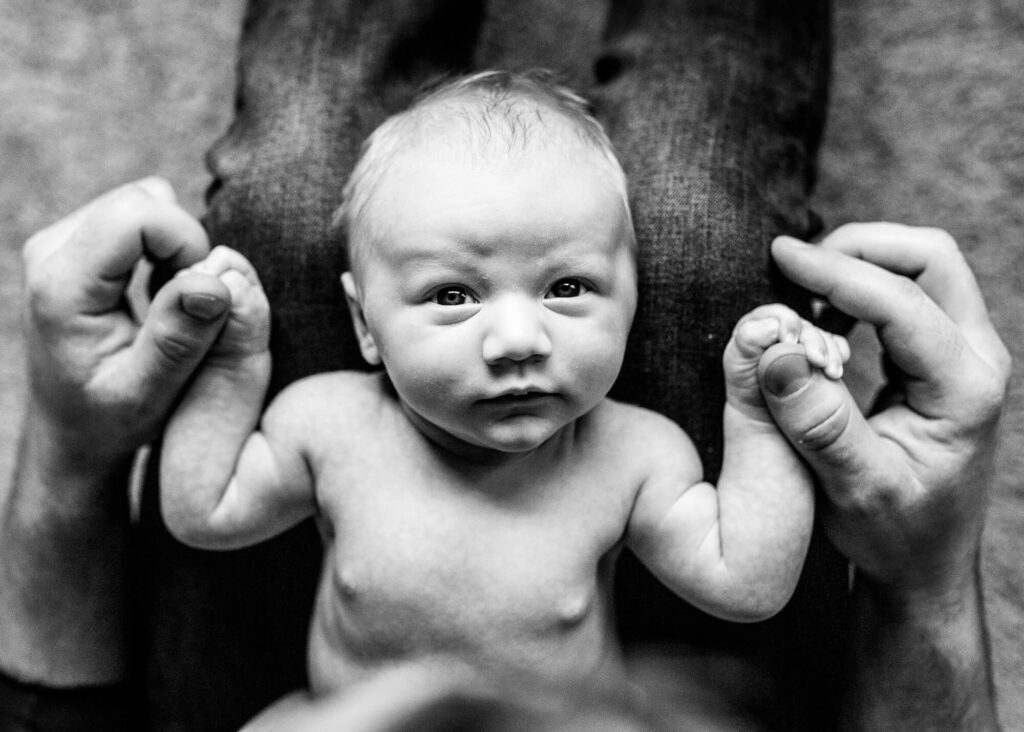 Black and white photo of newborn holding dad's hands in his lap. | Portland Midwifery 
