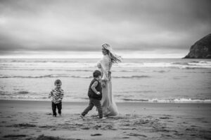 A black and white image of a mom in a long dress and two little boys playing at the Pacific City, Oregon beach. Things to do in Pacific City, Oregon, for Families.
