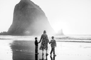 Black and white photo of a mother holding her two sons' hands in front of Haystack Rock in Cannon Beach, Oregon.