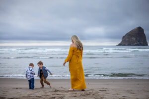Mom of two little boys wearing a yellow dress is playing on the beach in front of Haystack Rock in Pacific City, Oregon. 
