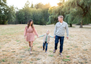 A family of three is walking in front of a large willow tree in a park near Portland, Oregon. Portland Photographer Mini Session.