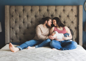 New parents look at one another as they hold their newborn son on a king bed. 