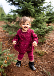 A baby girl in red holiday outfit smiling at a tree farm near Portland, Oregon. 