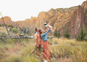 Family of three in the field at Smith Rock State Park playing with their baby at sunset for a family photo shoot. 