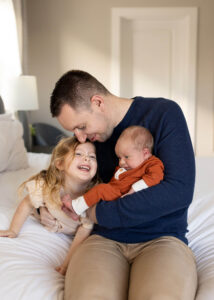 Dad holding newborn son with young daughter laughing by him. 