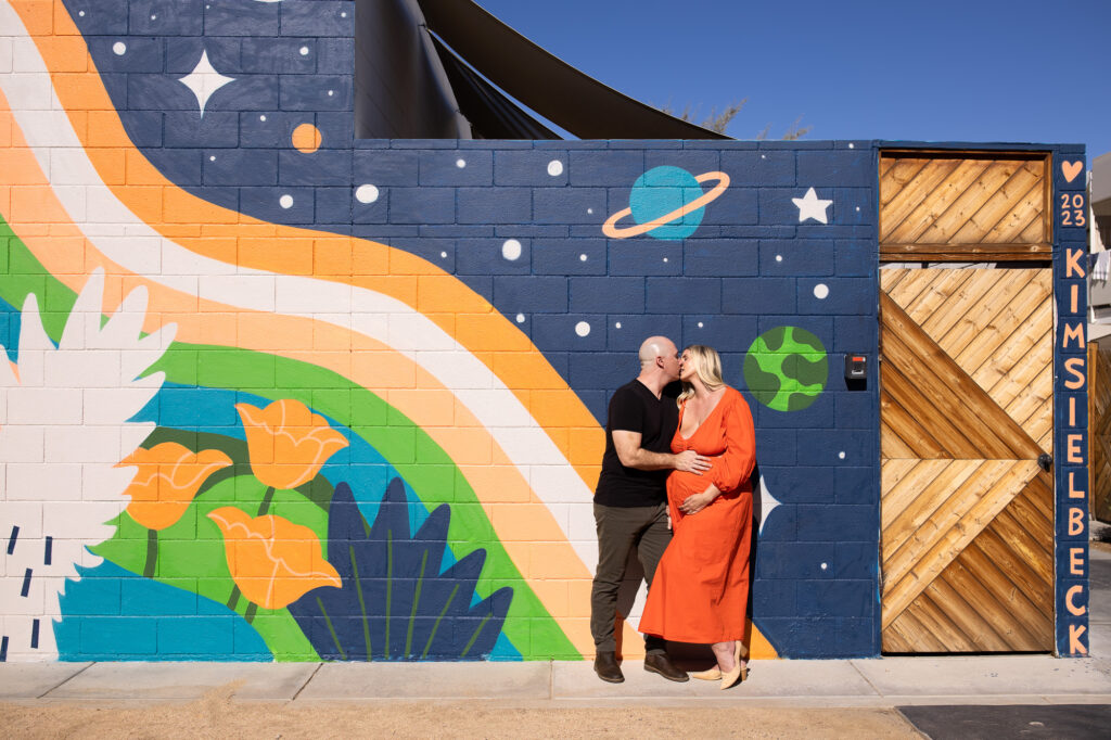 Couple stands in front of a colorful mural for a Palm Springs maternity photography session. 