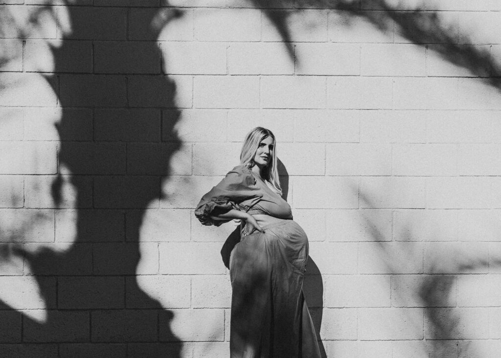 Black and white photo of a pregnant woman with a palm tree shadow on the white brick wall behind her. 