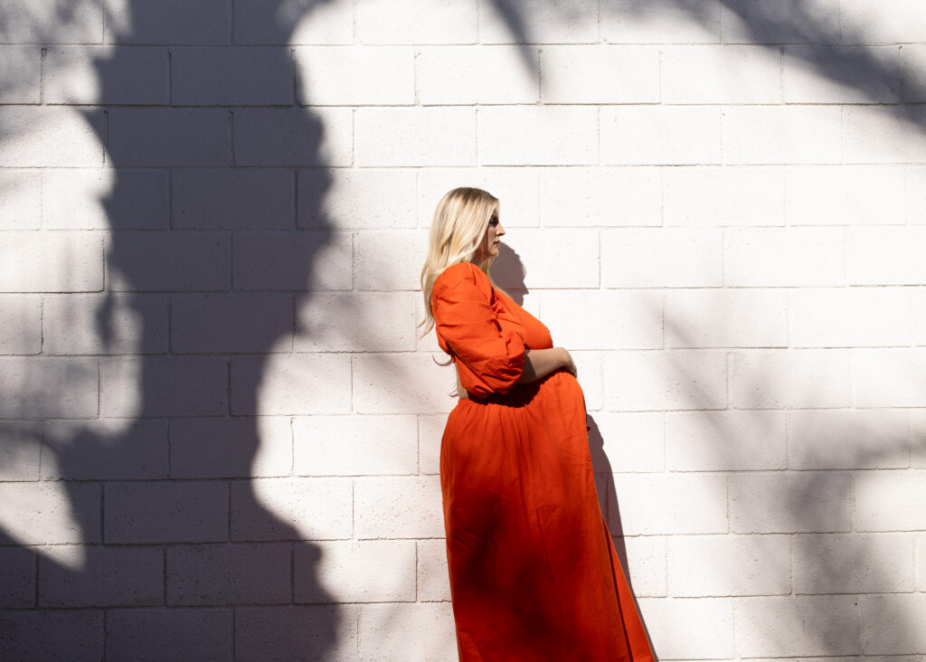 Pregnant woman in orange dress standing against a white brick wall for Palm Springs maternity photography session. 
