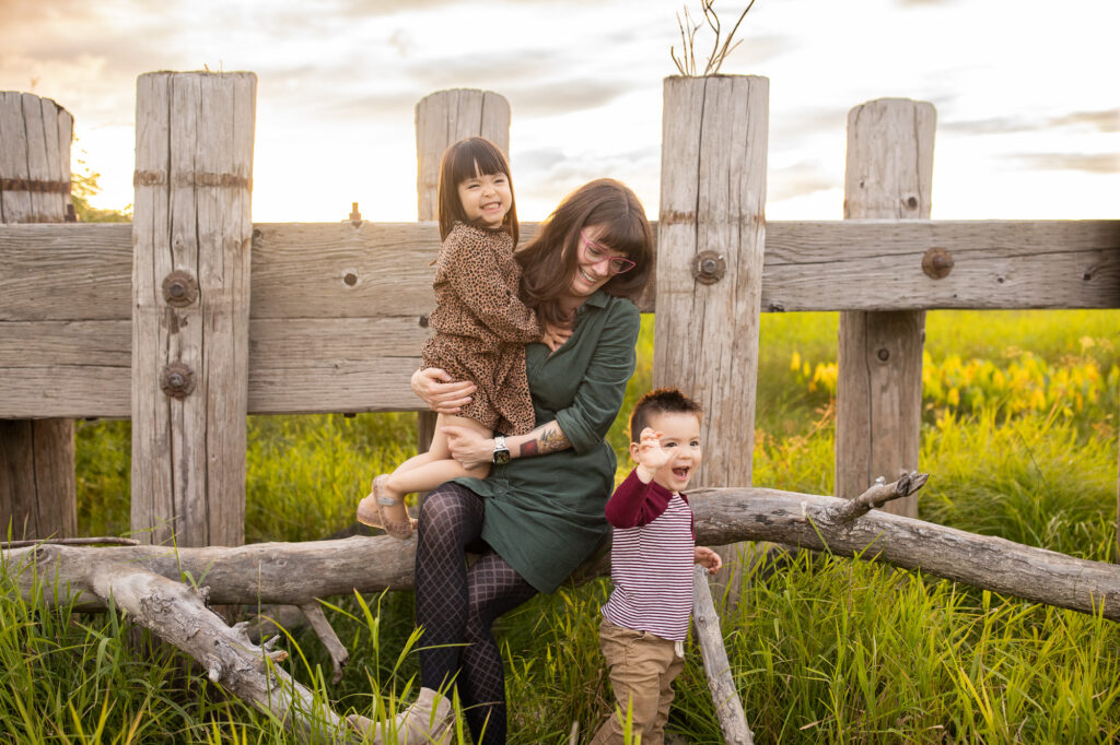Mother sits with two young kids against fence on a log with vibrant green grasses at sunset. 