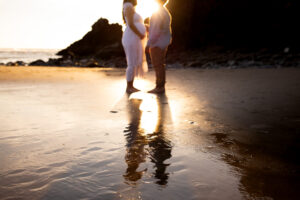 Silhouette of expecting parents holding hands with the sunlight reflecting on the sand — a dreamy idea for pregnancy photoshoots.