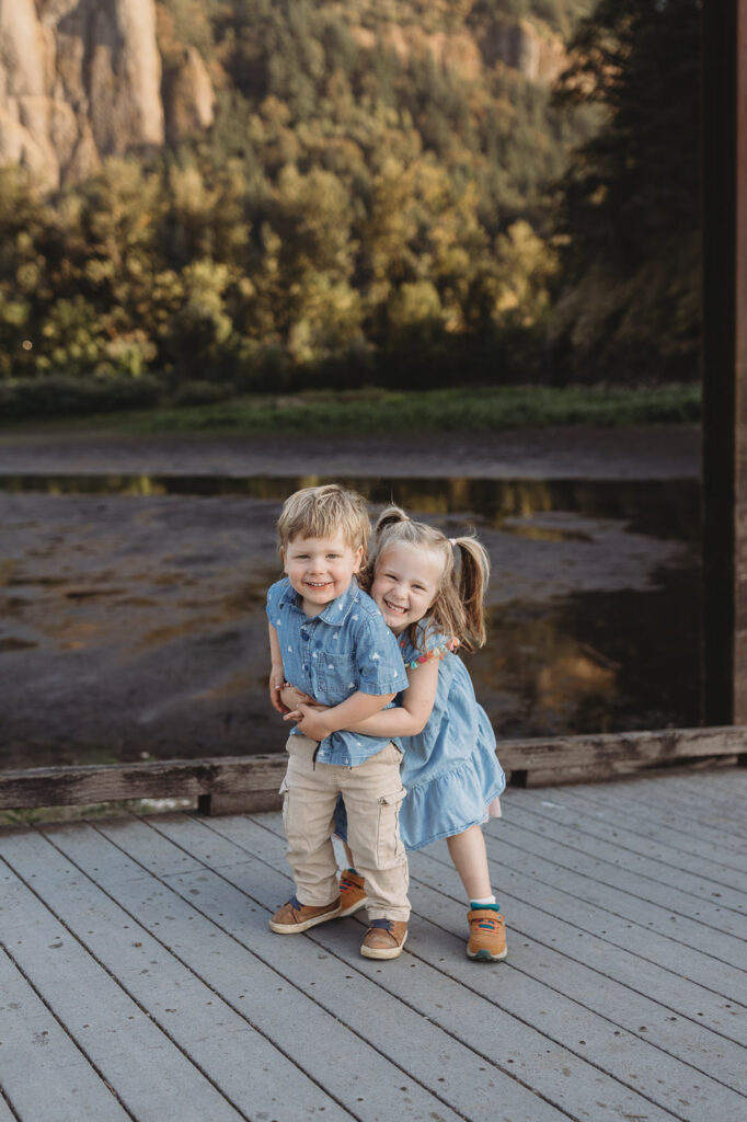 Toddler and his sibling hug on a dock at sunset in an adventure family photo shoot. 