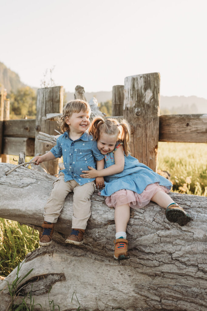 Siblings sit on a log on the river bed at sunset.